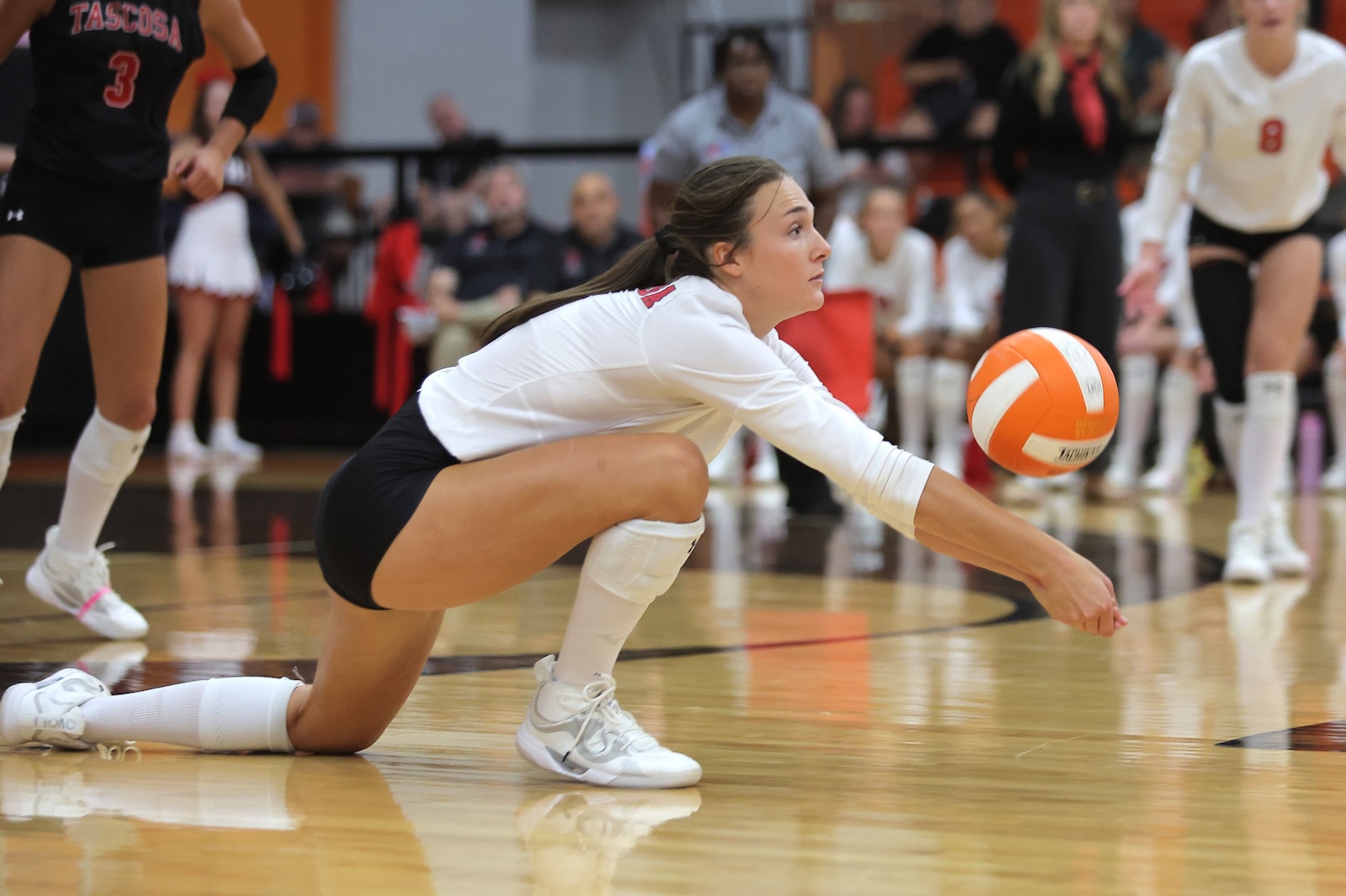 Female volleyball player in white jersey dives on the court, reaching for an orange-white ball near the floor.