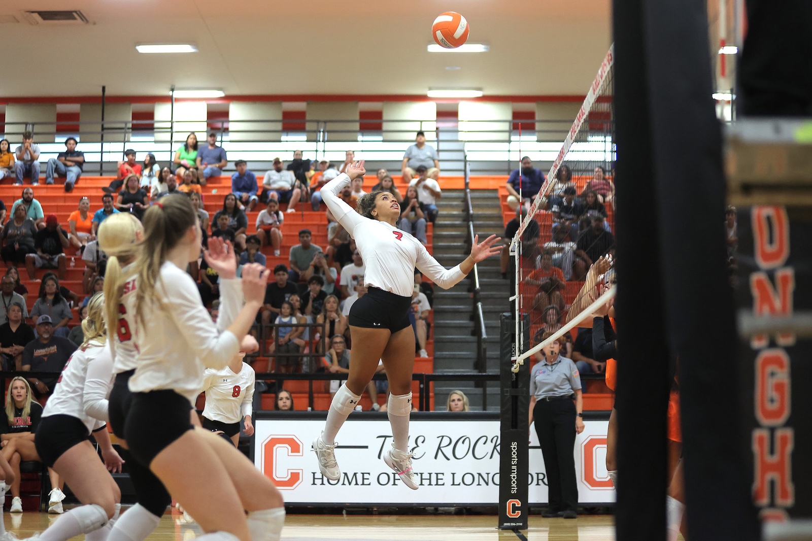 Volleyball player in a white jersey jumps near the net during a match as spectators watch from orange seats.