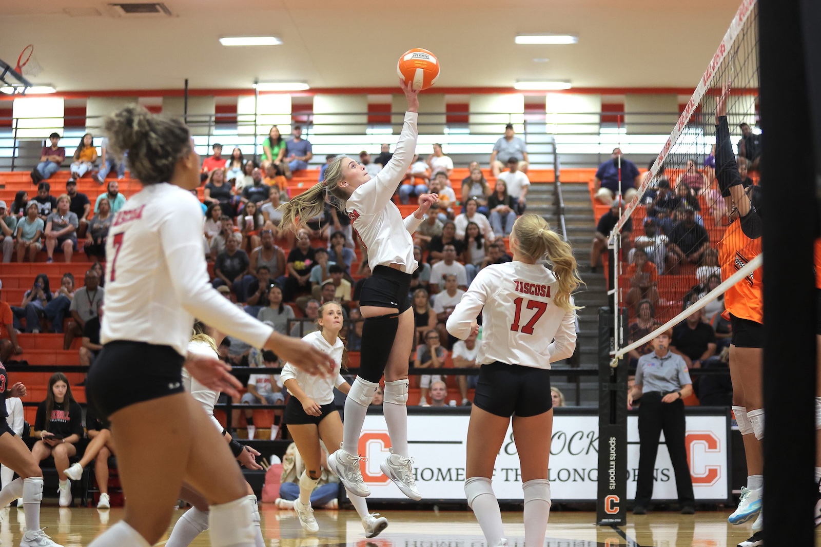 Female volleyball player in white jersey leaps to spike an orange ball over the net as teammates and a crowded gym observe the play.