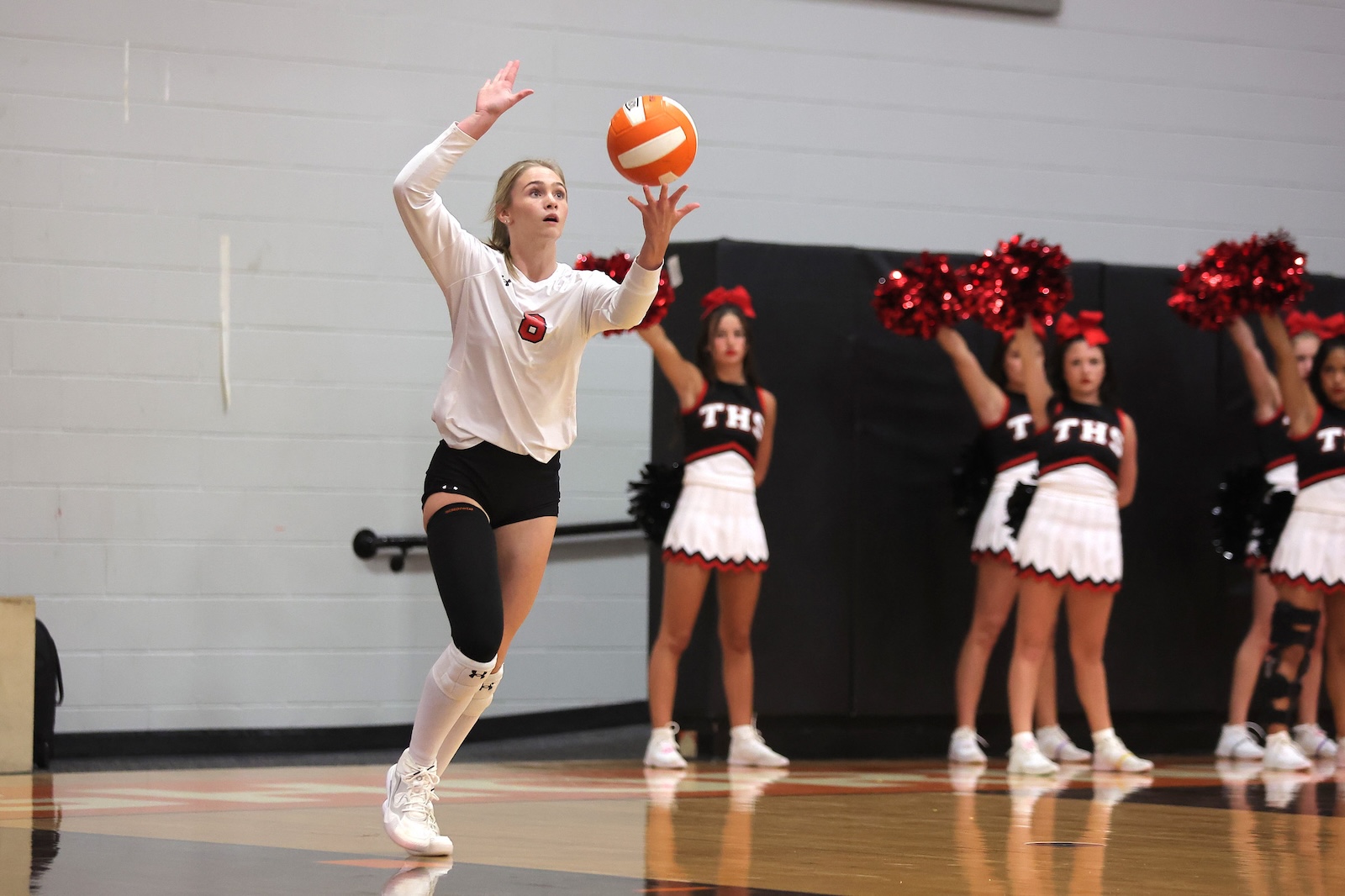 Female volleyball player in a white jersey (number 9) jumping to hit a volleyball in a gym, with cheerleaders in red and white uniforms in the background.
