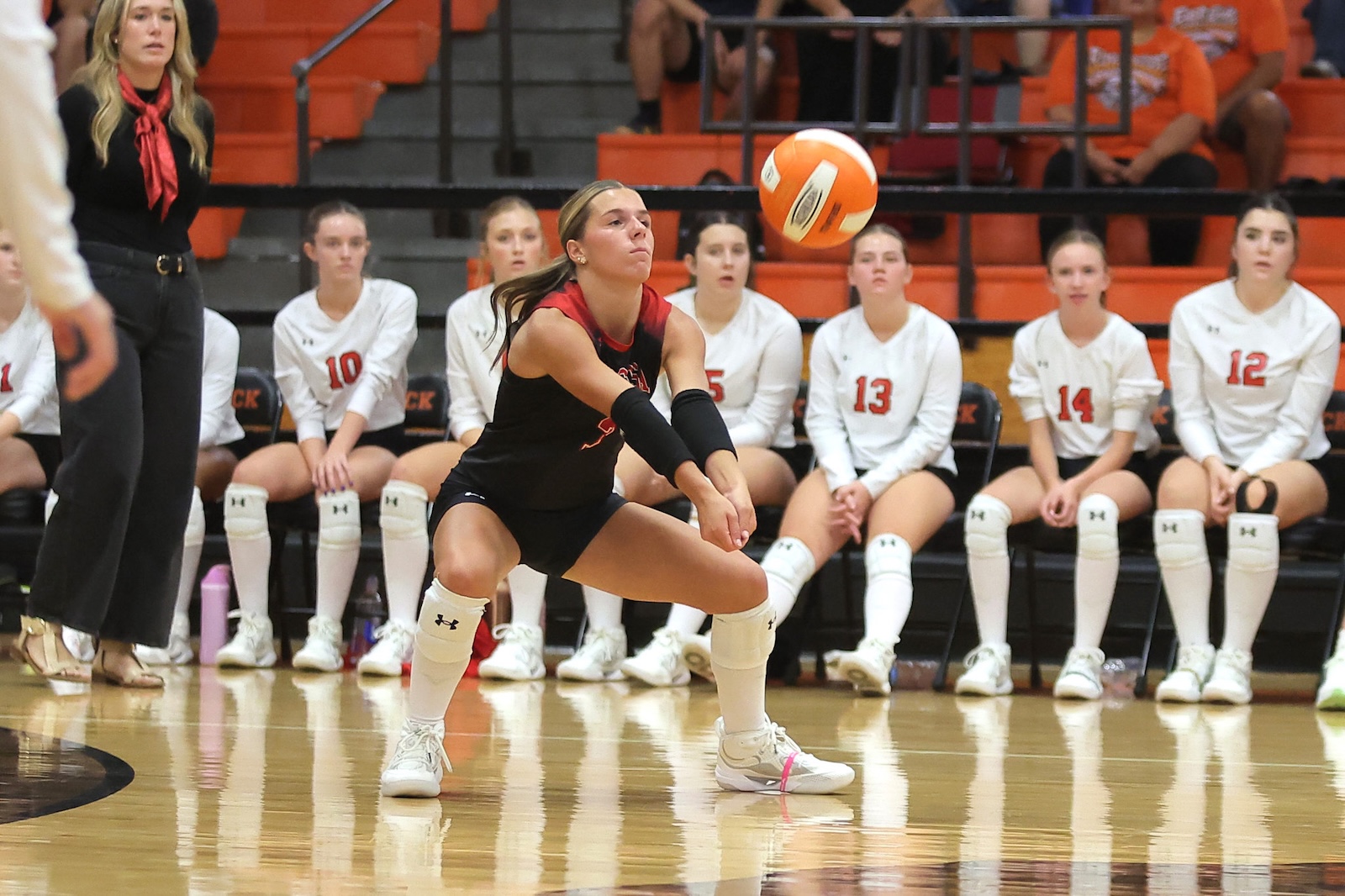 Female volleyball player in a black uniform dives to receive the ball, arms extended, as teammates in white sit on the bench watching in a gym.