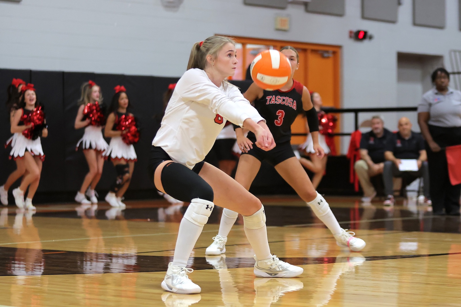 Female volleyball player in a white jersey and knee pads lunges to pass a spinning orange ball during a match, with cheerleaders and spectators in the background.