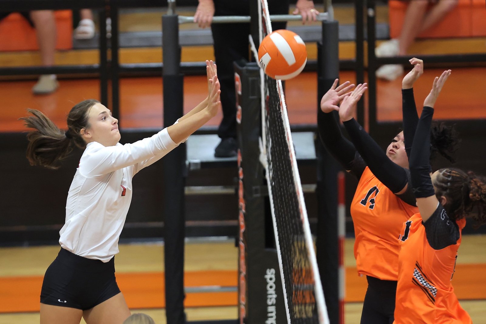 Female volleyball player in a white jersey dives toward the net to block as a volleyball hovers between blockers and attackers in an orange-jersey match in a gym.