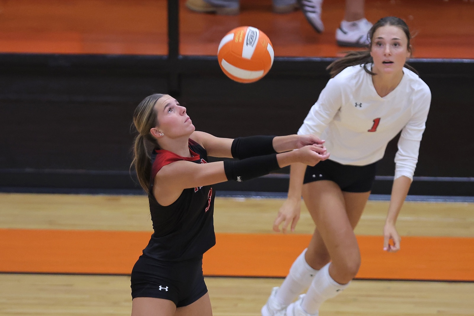 Two female volleyball players reach for an orange-and-white ball during a match; one in black extends for a pass while a teammate in white watches nearby.
