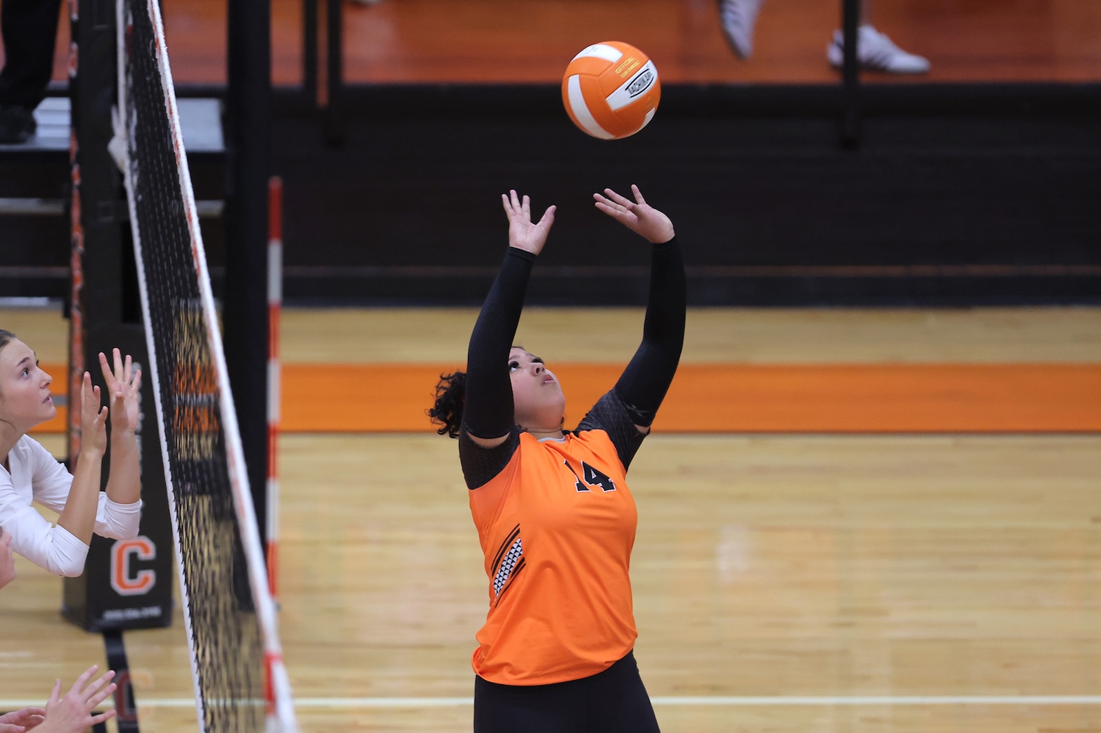 Female volleyball player in orange jersey sets the ball over the net during a match, with a teammate watching from the left in a gymnasium.