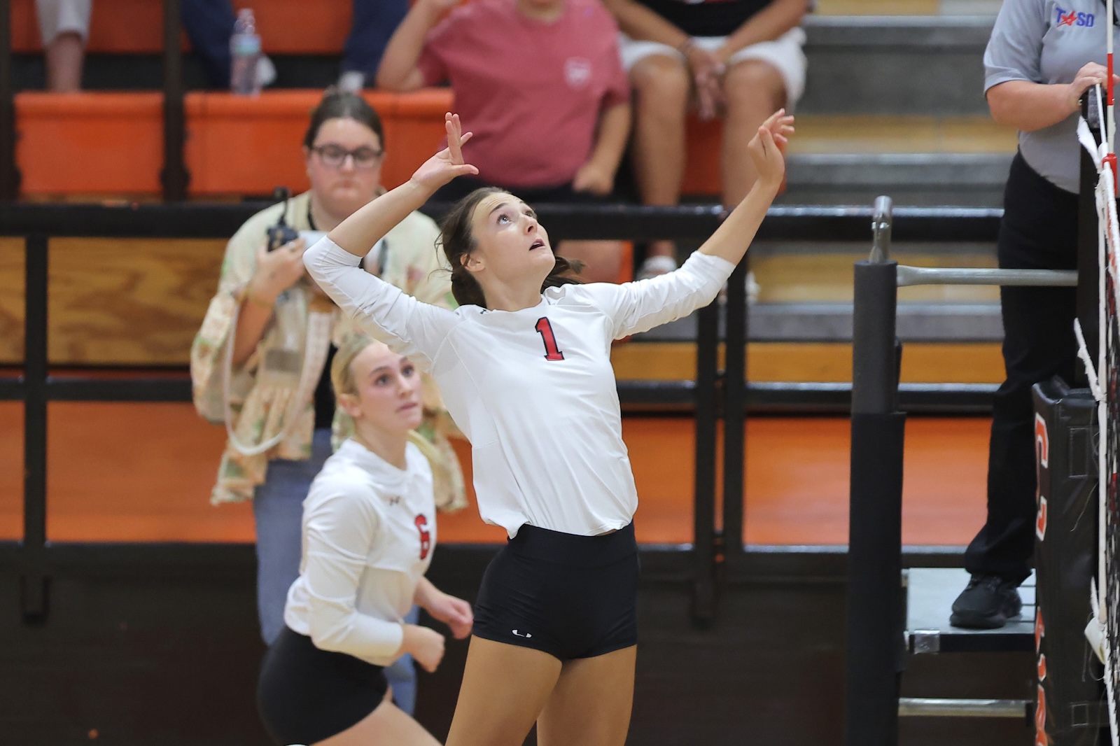 Volleyball player wearing #1 in white jersey jumps with arms raised at the net during a match, a teammate watches nearby.