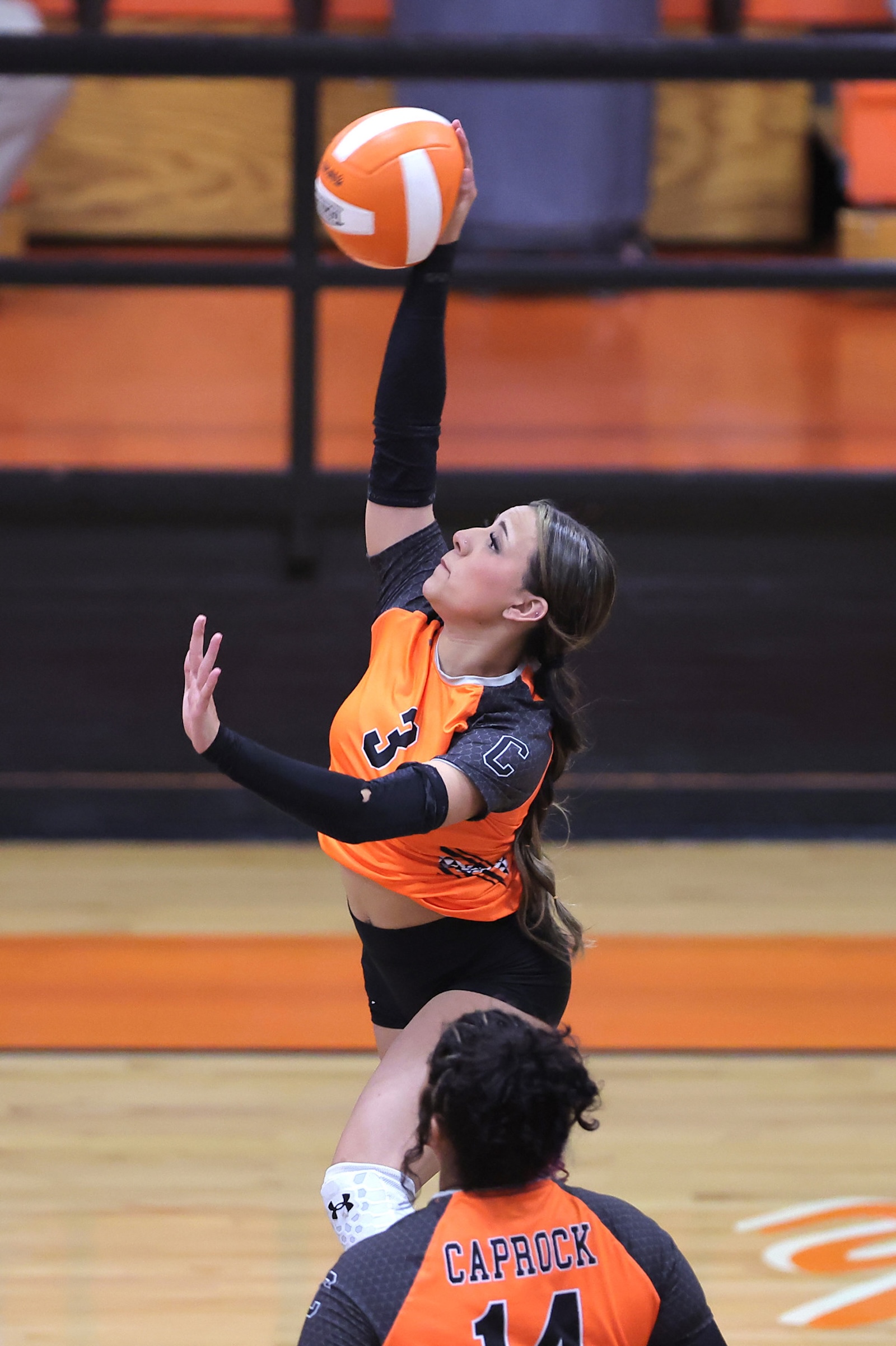 Female volleyball player in orange jersey jumps to spike the ball, arm extended above her head in an indoor gym.