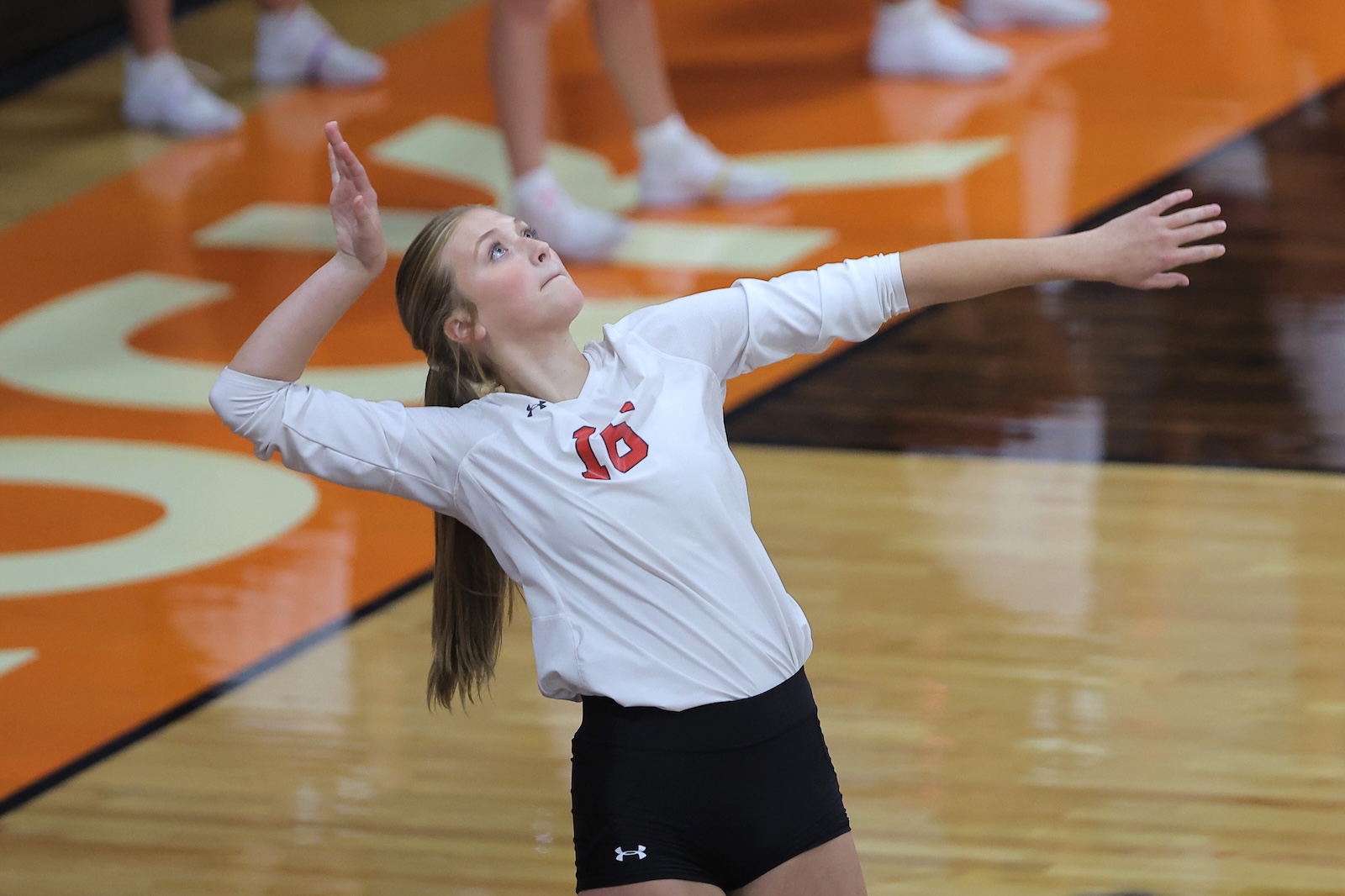 Young female volleyball player in a white jersey (number 16) extending her arms in a serving motion on a gym floor with orange and white markings.