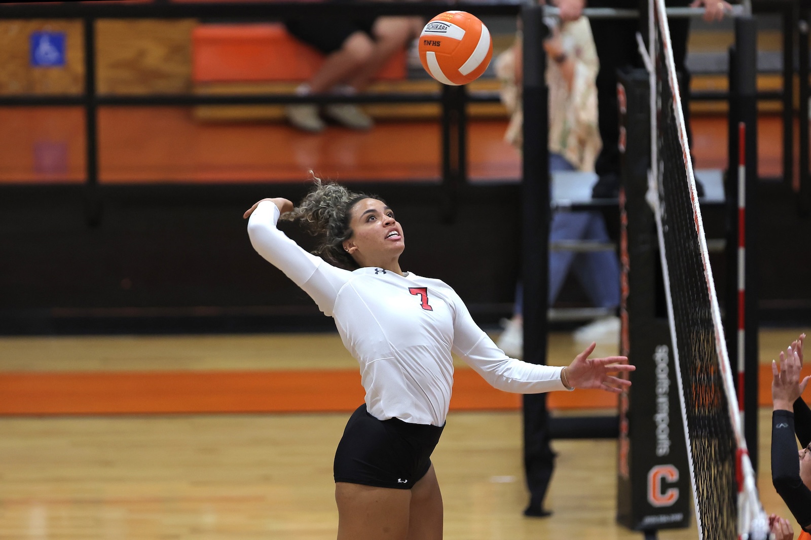 Female volleyball player in white jersey (number 7) spikes the ball near the net during a match in a gymnasium.