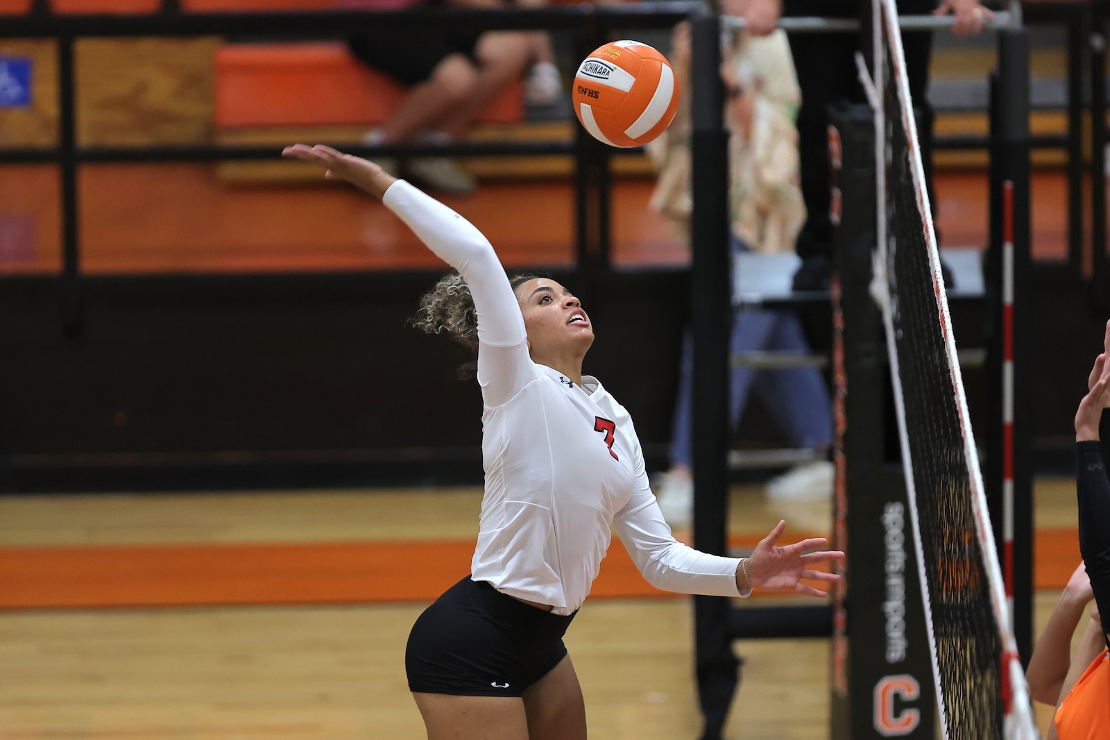 Female volleyball player in a white long-sleeve jersey (number 7) spikes the ball over the net during a match in a gymnasium