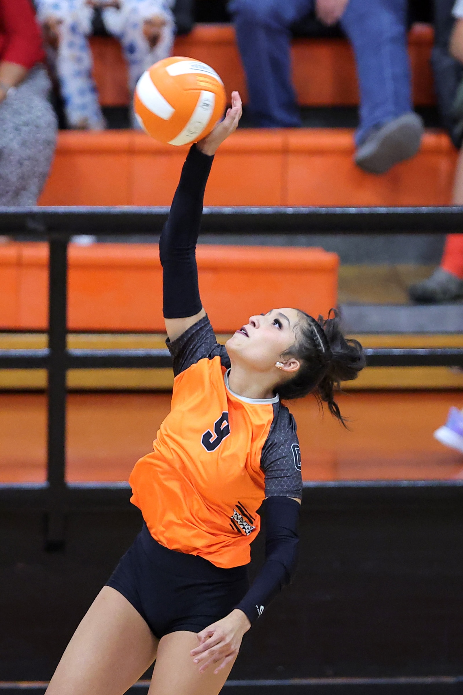Female volleyball player in an orange jersey swings her arm to spike a ball during a game in a gym with orange bleachers behind her.