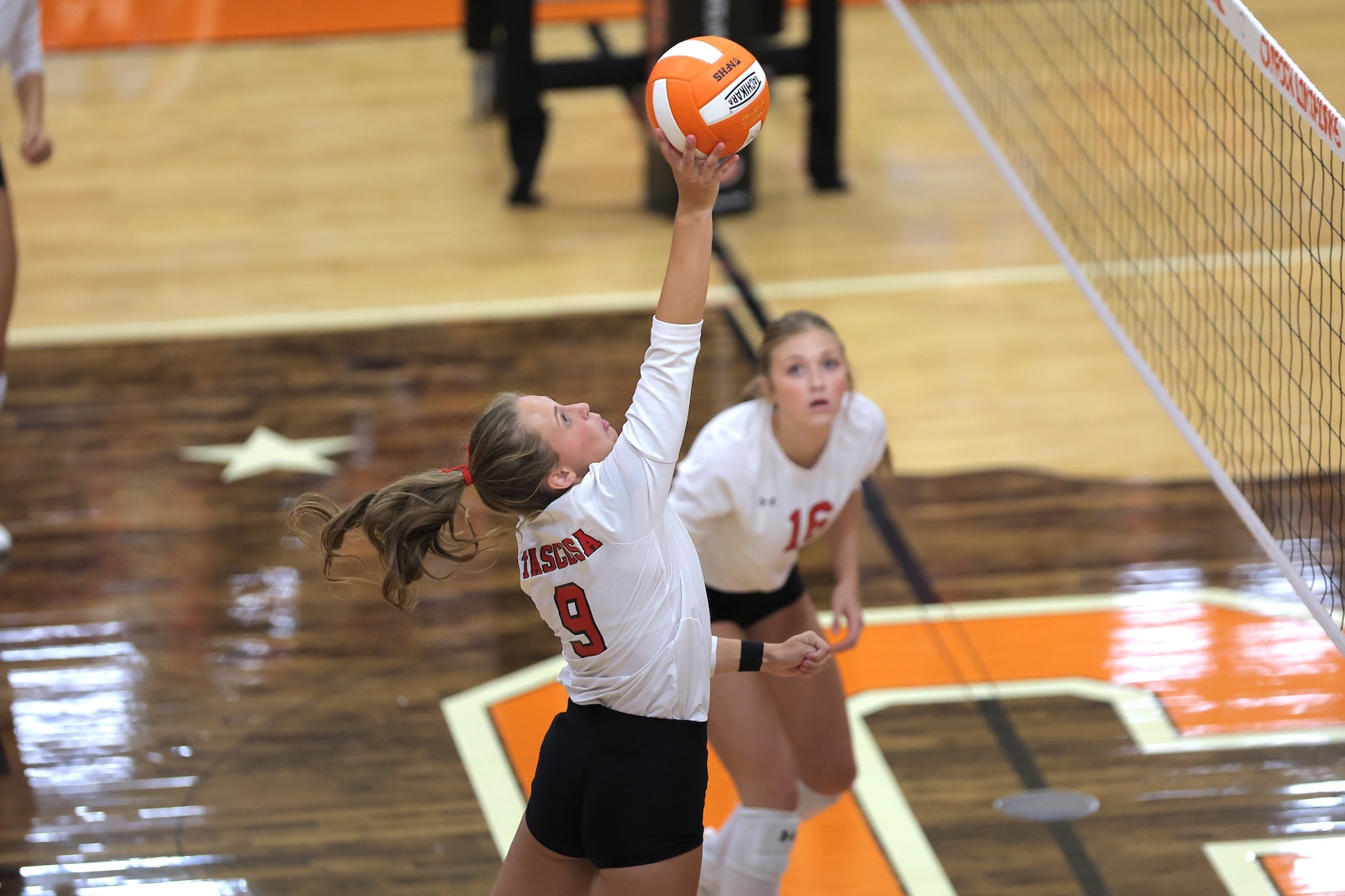 Volleyball player in white jersey (#9) jumps with arm raised to hit the orange ball over the net while a teammate watches nearby in a gym.