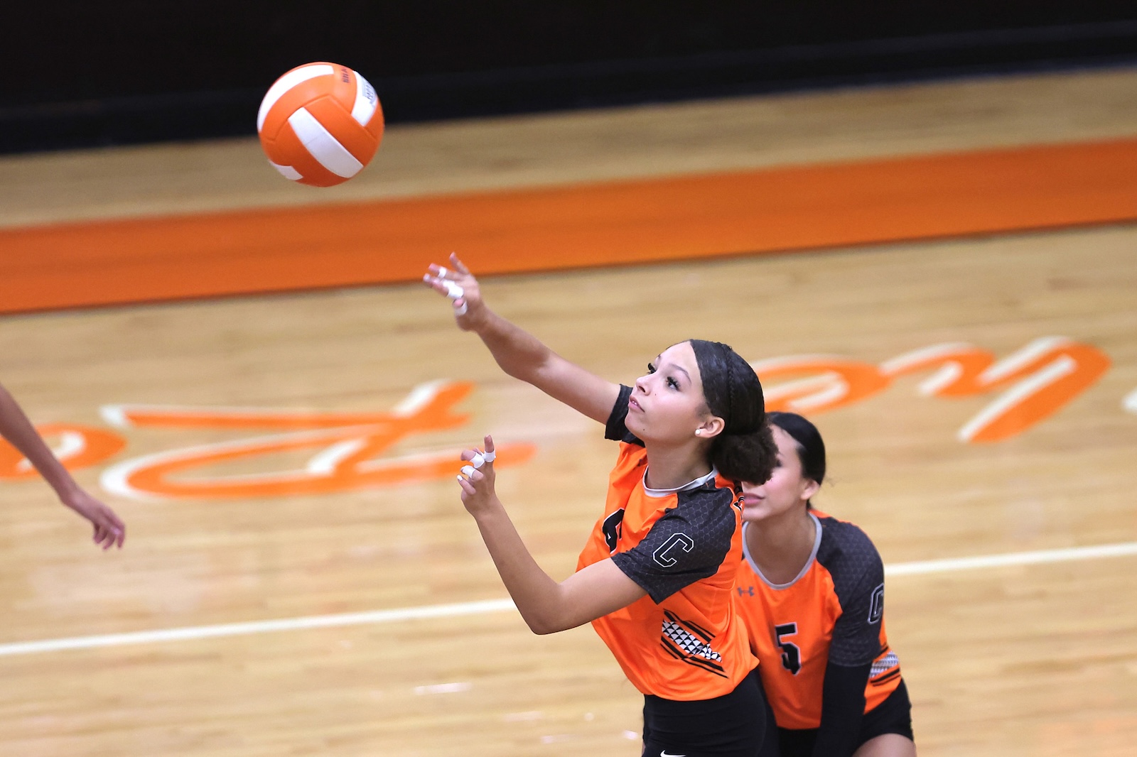 Two female volleyball players in orange uniforms reach for an orange-and-white ball during a fast-paced indoor match on a polished wood court.