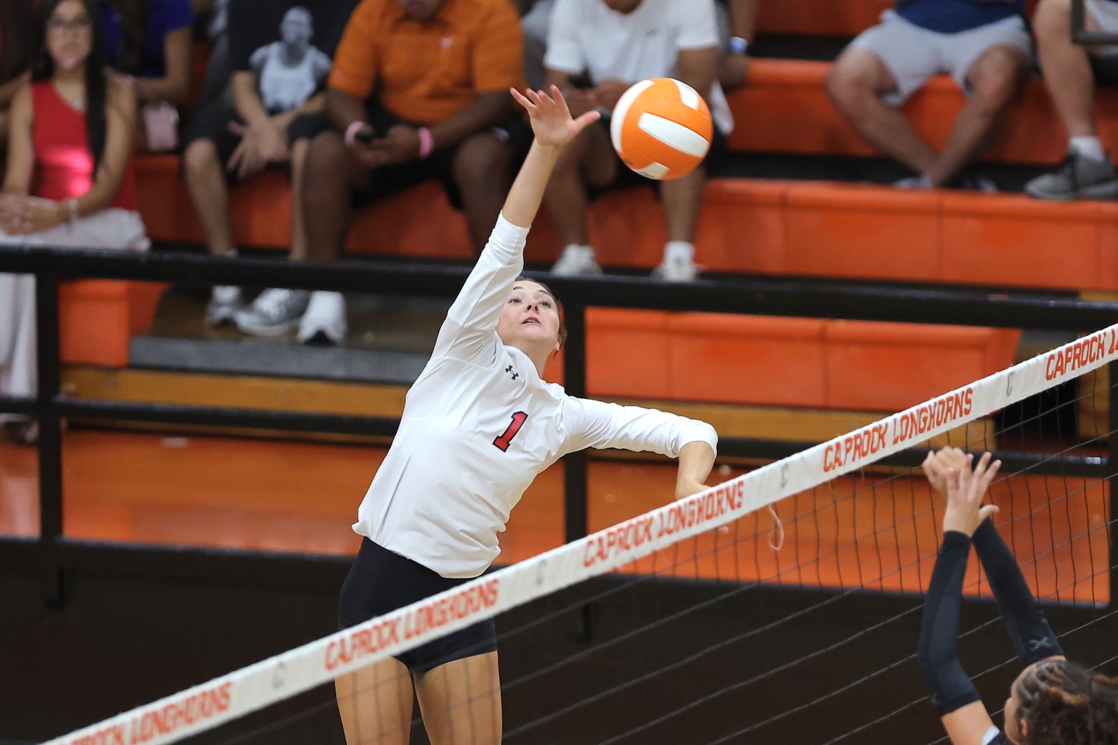 Female volleyball player in a white jersey with #1, leaping to hit the ball over the net as spectators watch from orange bleachers.