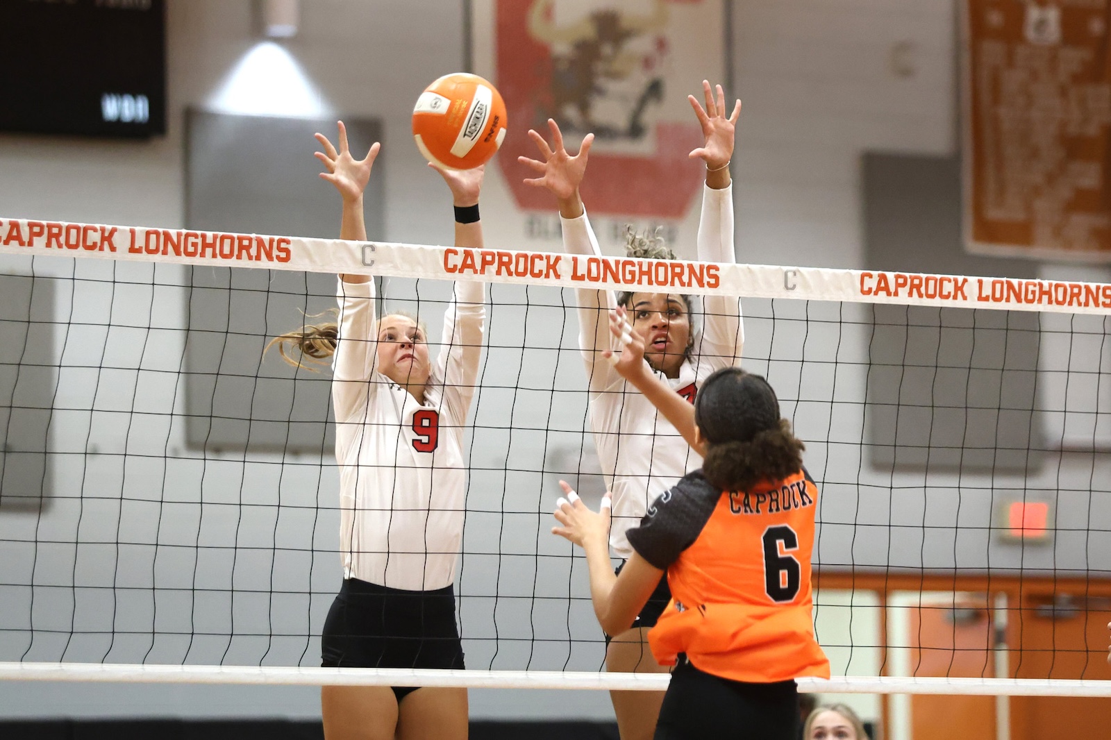 Two players in white jerseys jump to block a volley at the net while an opponent in orange approaches.