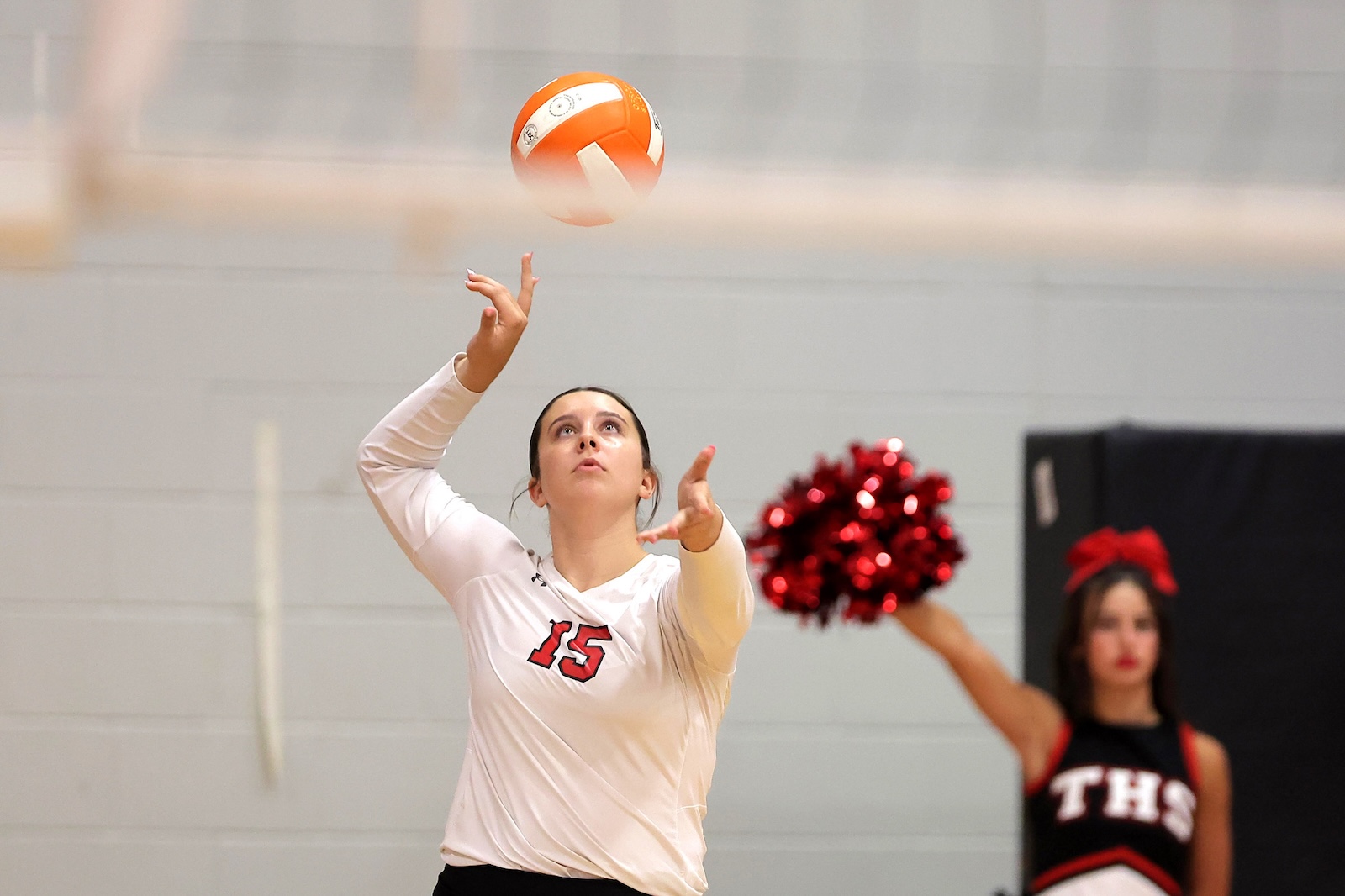 Volleyball player in a white jersey number 15 prepares to set the ball overhead in a gym, with a cheerleader in the background.
