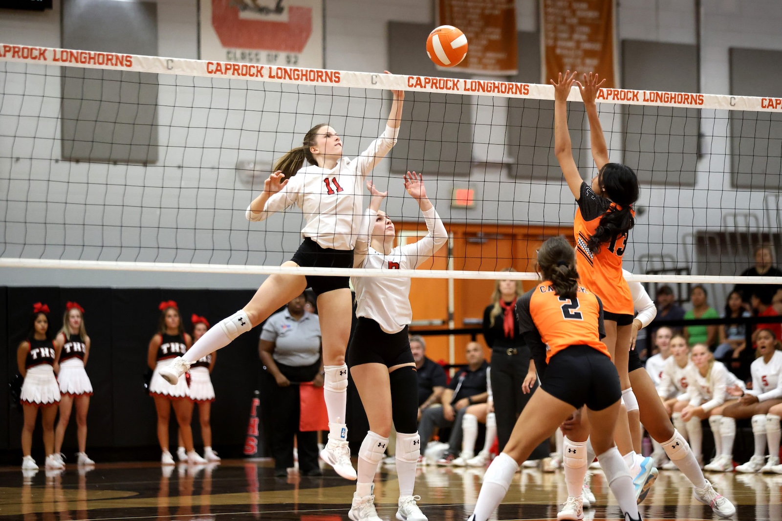 Volleyball game in a high school gym; white-team player (11) jumps to spike the ball over the net as an orange-team blocker reaches up.