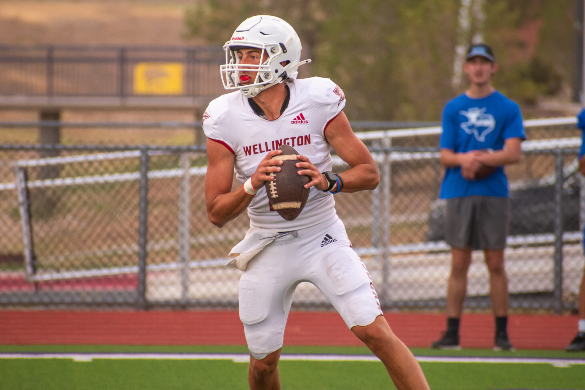 DSC_0344 | Press Pass Sports Wellington football player in a white uniform and helmet runs with the football, chasing downfield, with a spectator in blue in the background.
