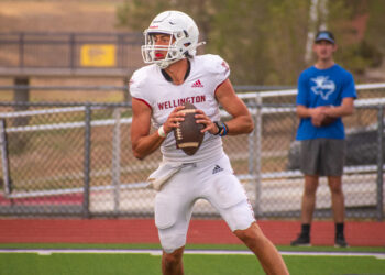 DSC_0344 | Press Pass Sports Wellington football player in a white uniform and helmet runs with the football, chasing downfield, with a spectator in blue in the background.