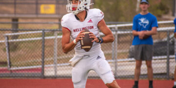 Wellington football player in a white uniform and helmet runs with the football, chasing downfield, with a spectator in blue in the background.