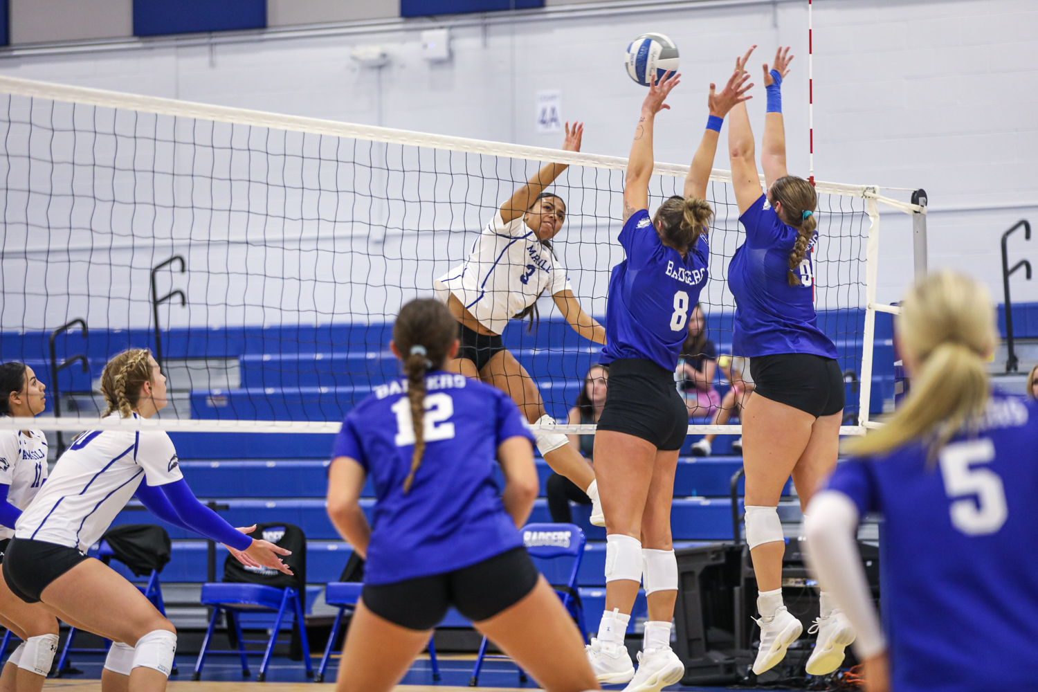 VB 4 | Press Pass Sports Volleyball players jump at the net as two blue-team blockers attempt to stop a white-team attack, ball in midair.