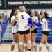 Group of female volleyball players in white jerseys huddling and celebrating on a gym court.