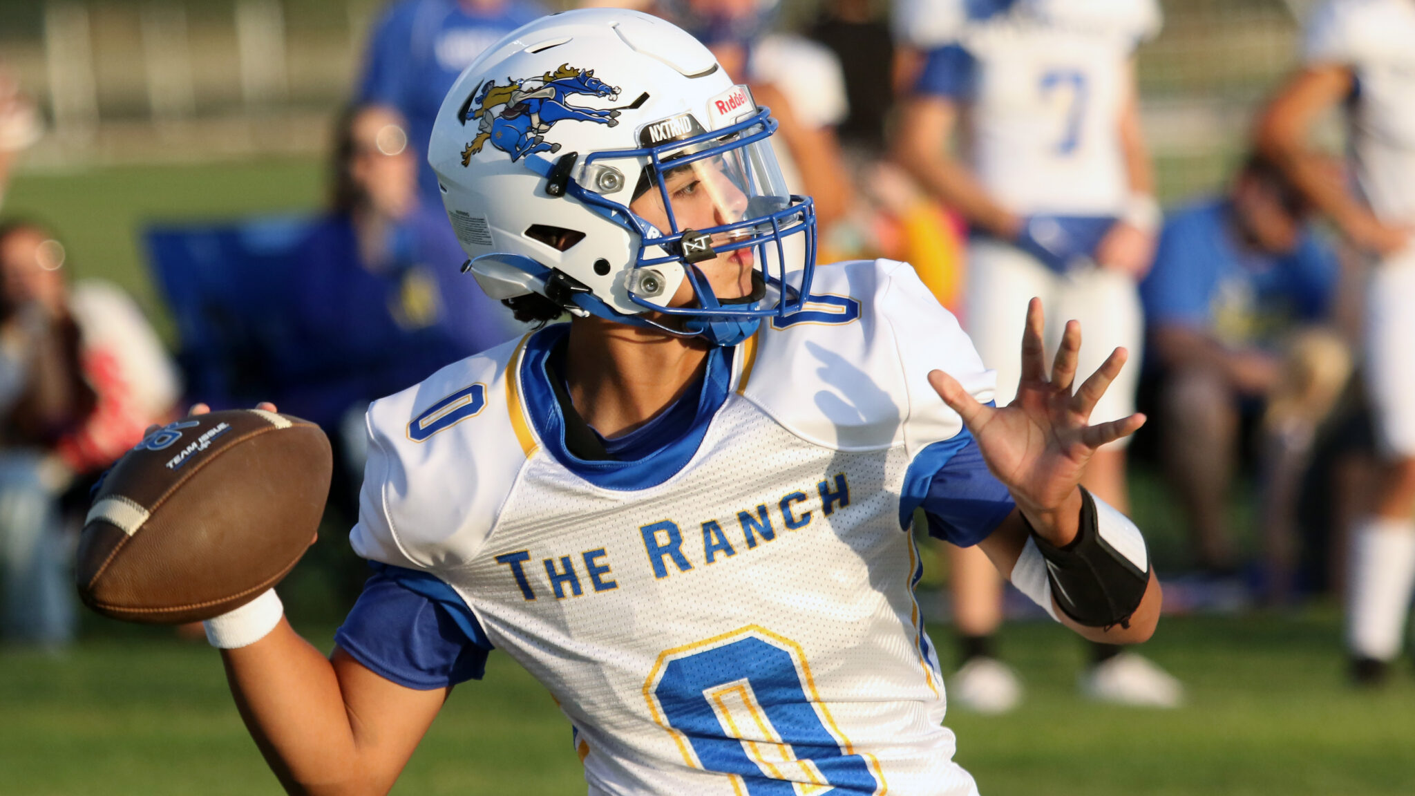 BoysRanch-SJCA17 2 | Press Pass Sports Football player in white and blue jersey with a helmet, holding a football and running during a game as blurred spectators watch in the background.