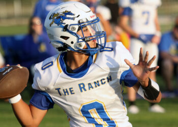 BoysRanch-SJCA17 2 | Press Pass Sports Football player in white and blue jersey with a helmet, holding a football and running during a game as blurred spectators watch in the background.