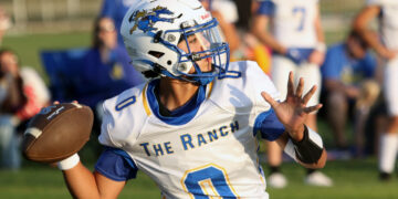 Football player in white and blue jersey with a helmet, holding a football and running during a game as blurred spectators watch in the background.