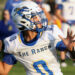 BoysRanch-SJCA17 2 | Press Pass Sports Football player in white and blue jersey with a helmet, holding a football and running during a game as blurred spectators watch in the background.