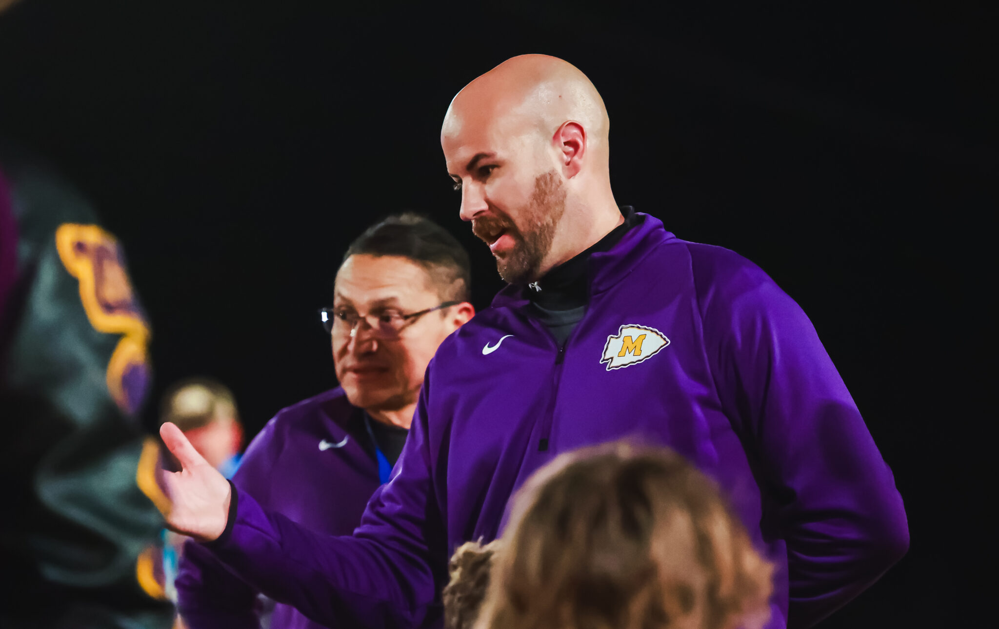 MY4A4934 1 | Press Pass Sports Bald man in a purple jacket speaking to a group at a nighttime event, gesturing with his hand.