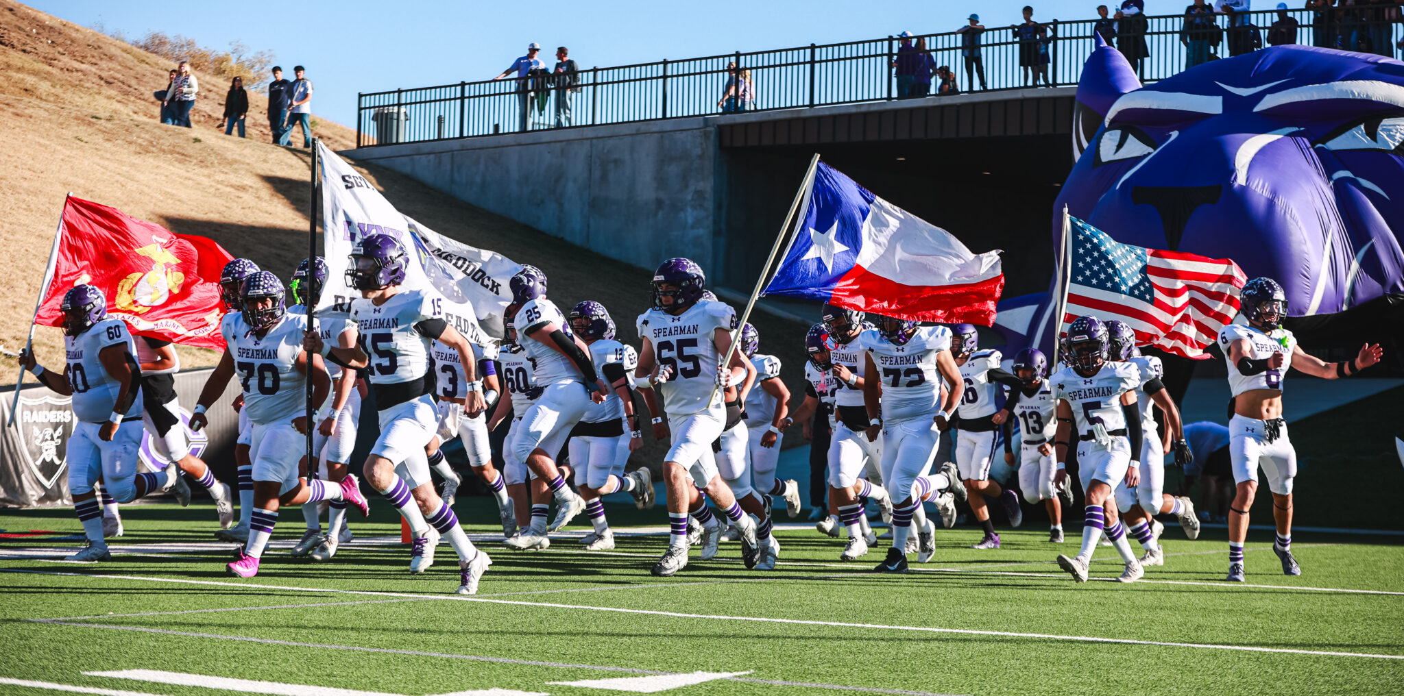 MY4A4954 | Press Pass Sports Spearman football players in white uniforms run onto the field carrying flags (Texas, USA) as fans watch from the stands above the tunnel area.