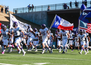 MY4A4954 | Press Pass Sports Spearman football players in white uniforms run onto the field carrying flags (Texas, USA) as fans watch from the stands above the tunnel area.