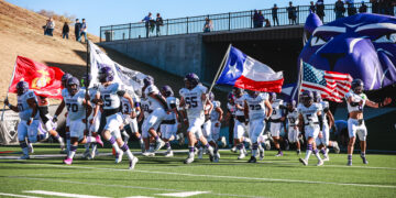 Spearman football players in white uniforms run onto the field carrying flags (Texas, USA) as fans watch from the stands above the tunnel area.