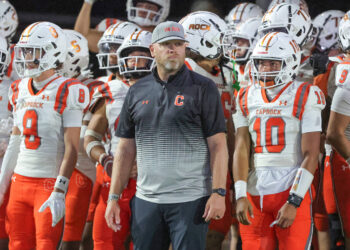 20250918-Caprock-Canyon-FB-0016 | Press Pass Sports Coach wearing a gray cap labeled 'THE ROCK' stands in front of Caprock football players in white and orange uniforms.