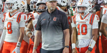 Coach wearing a gray cap labeled 'THE ROCK' stands in front of Caprock football players in white and orange uniforms.