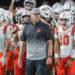 20250918-Caprock-Canyon-FB-0016 | Press Pass Sports Coach wearing a gray cap labeled 'THE ROCK' stands in front of Caprock football players in white and orange uniforms.