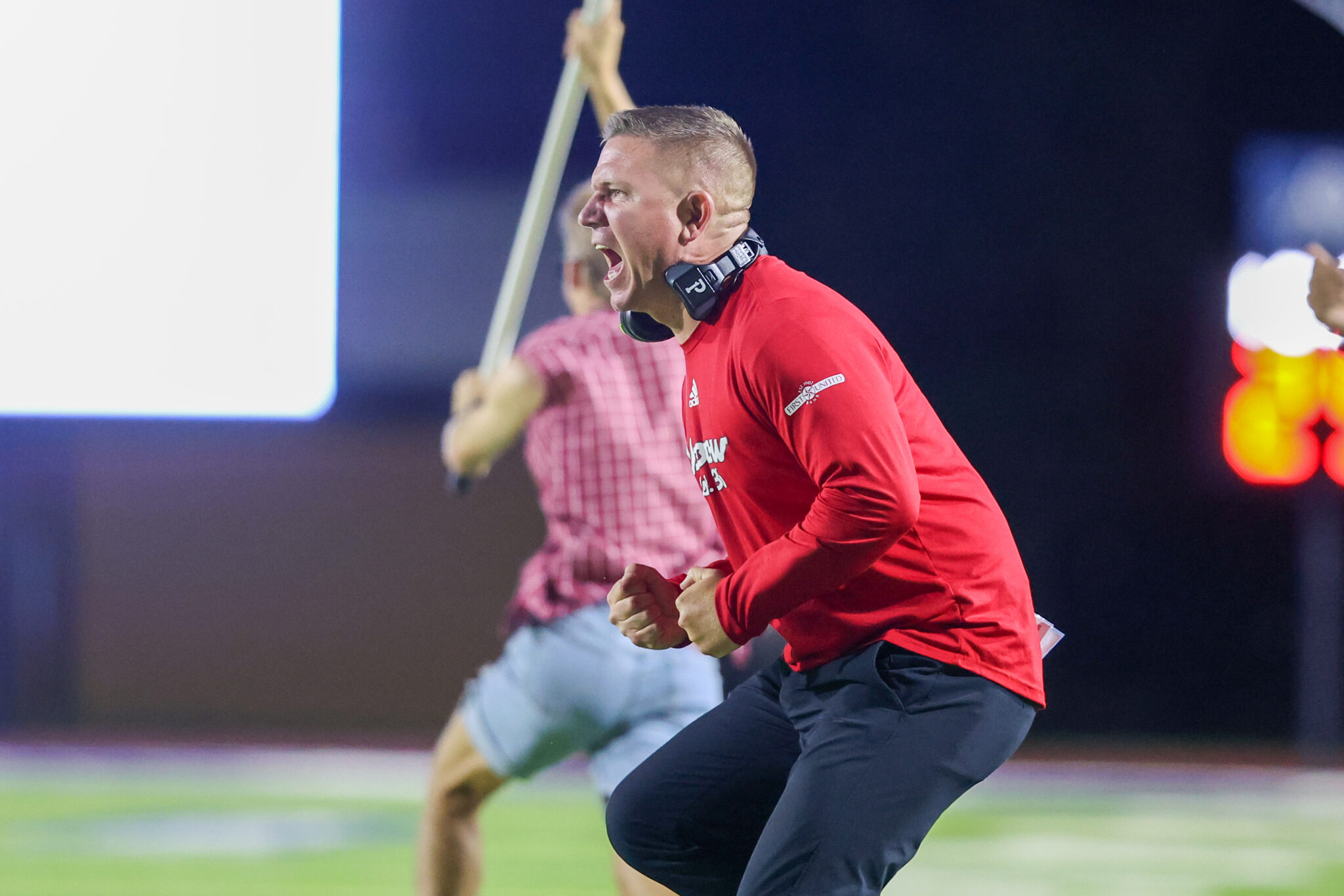 Seabolt Chase-6774 | Press Pass Sports Man in a red sports shirt runs forward, fists clenched in celebration during a night game on a lit field with an out-of-focus teammate behind him.