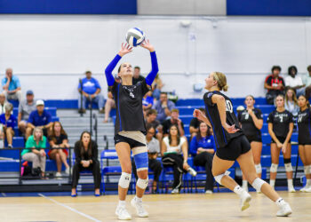 Volleyball players in black uniforms at net, one setter raising the ball for a teammate as spectators watch from blue bleachers.