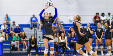 Volleyball players in black uniforms at net, one setter raising the ball for a teammate as spectators watch from blue bleachers.