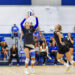 Volleyball players in black uniforms at net, one setter raising the ball for a teammate as spectators watch from blue bleachers.