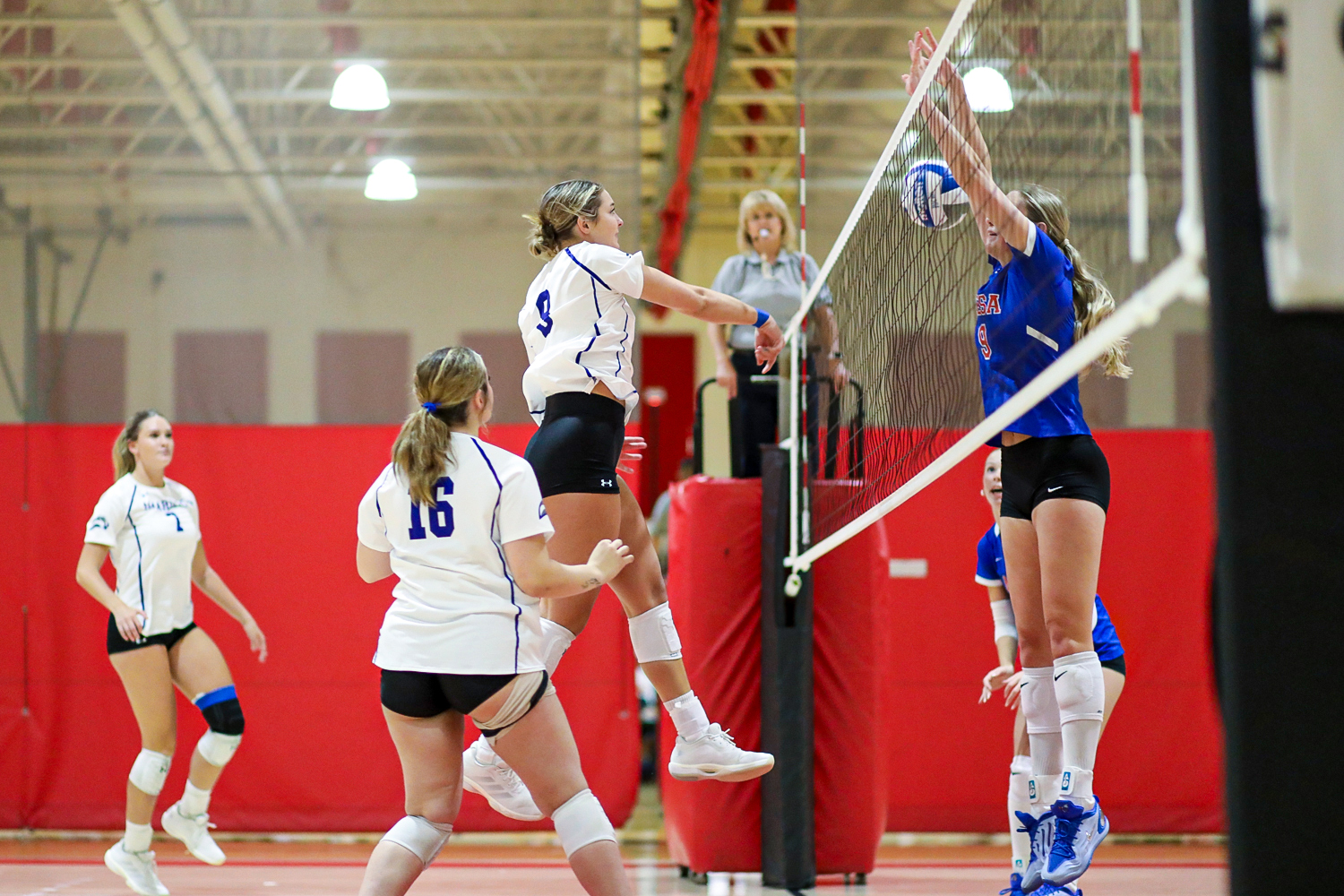 NMMI 3 | Press Pass Sports Two white-jersey players jump to block a blue-team spike at the net during a volleyball match.