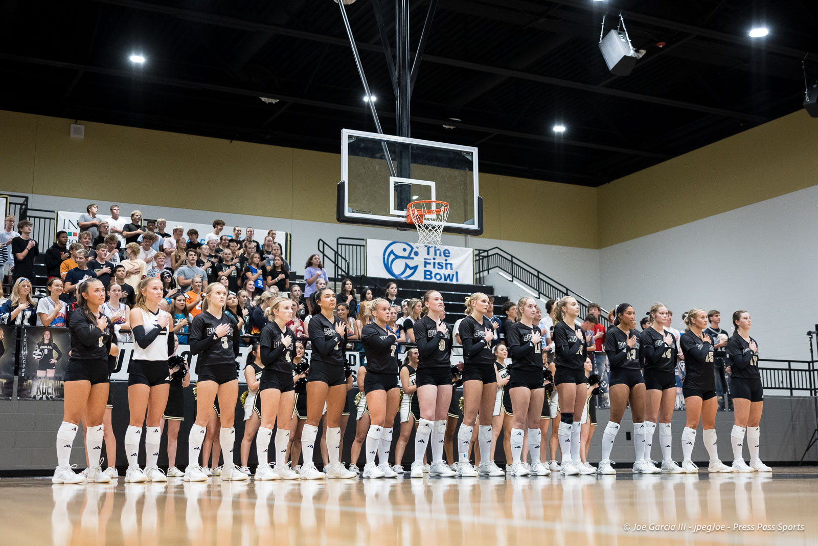Girls' volleyball team stands in a line on a gym floor with hands over hearts as fans watch from the bleachers behind them on a banner-adorned wall.
