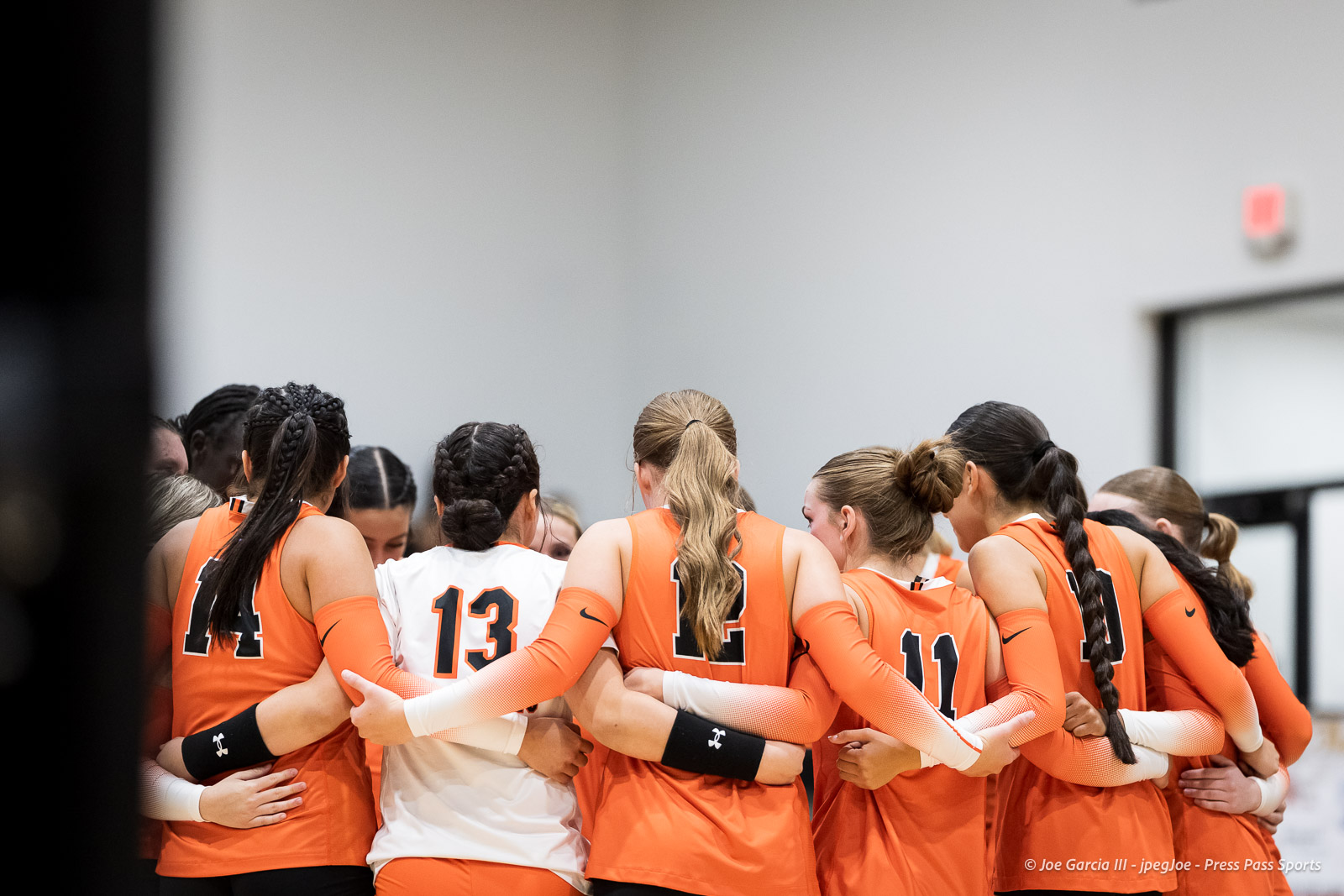 Women’s volleyball team in orange uniforms huddling with arms around each other.