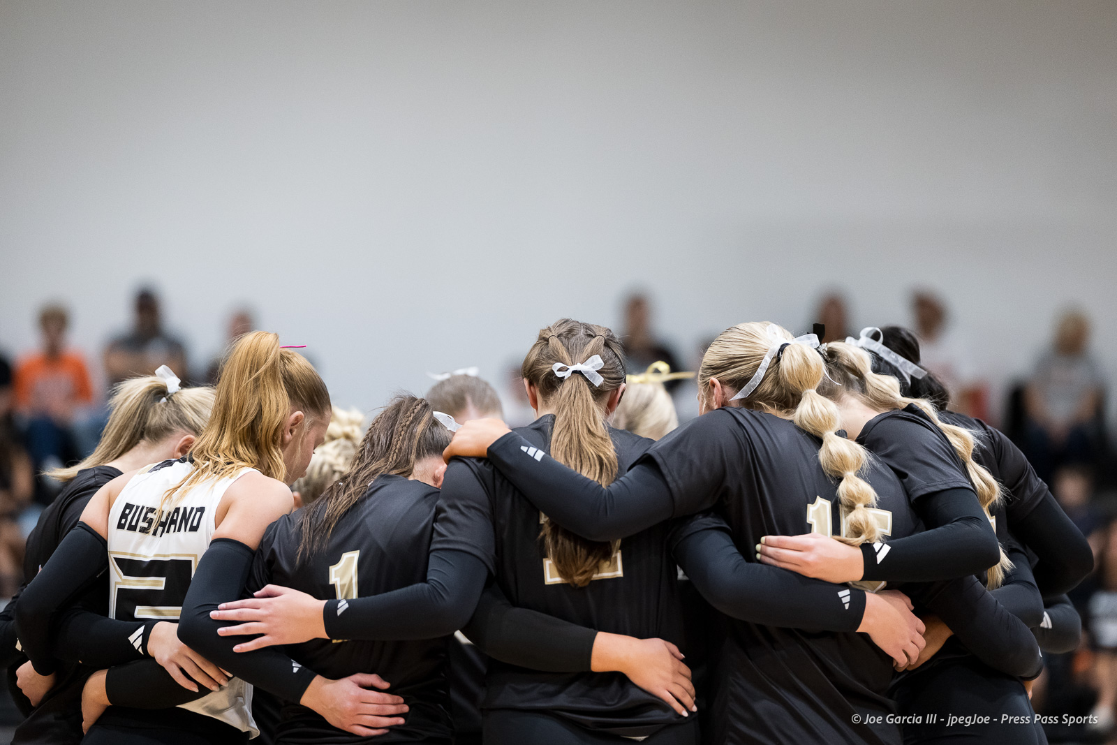 Girls' volleyball team huddled together with arms around each other before a game on the court.