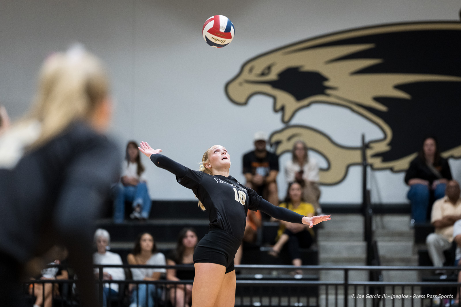 Female volleyball player in a black jersey #10 prepares to serve, ball in midair, with spectators watching from the bleachers and a large mascot mural in the background.