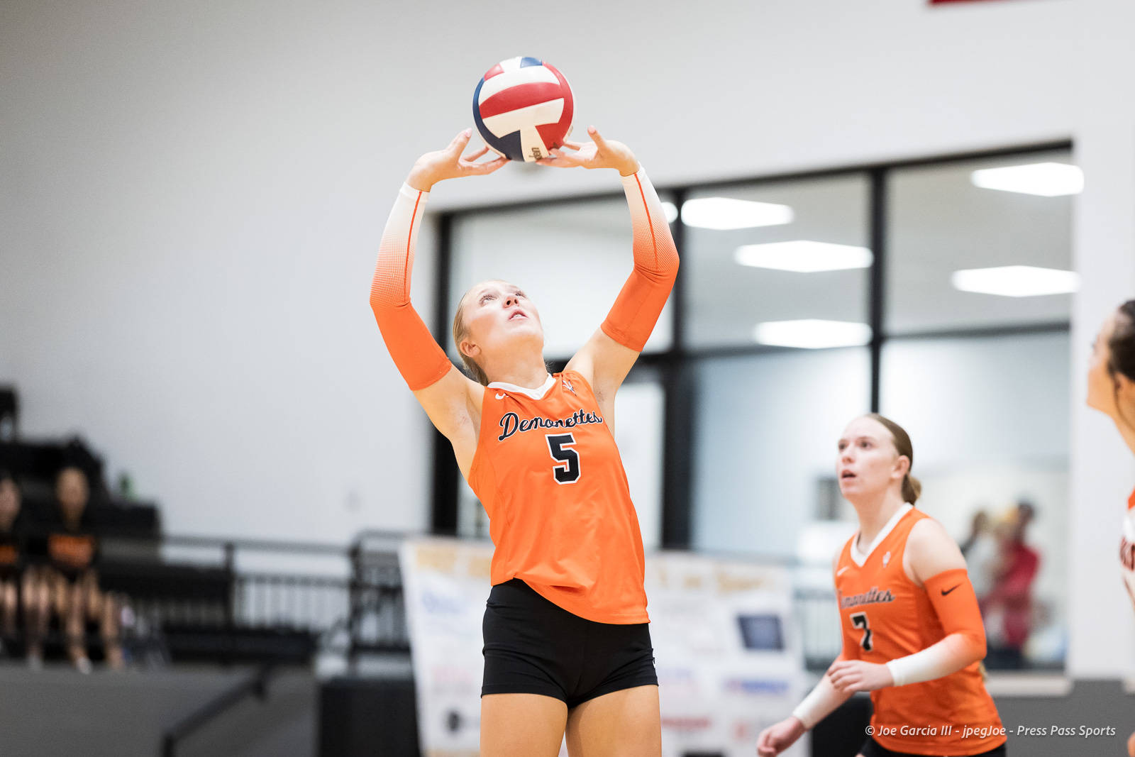 Female volleyball player in orange jersey #5 raises arms to set the ball during a match.