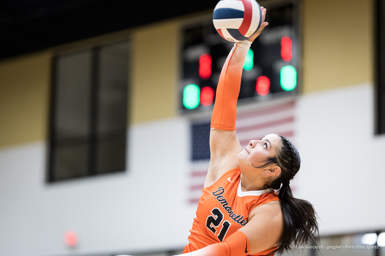Female volleyball player in an orange jersey reaching up to spike the ball in a gymnasium.