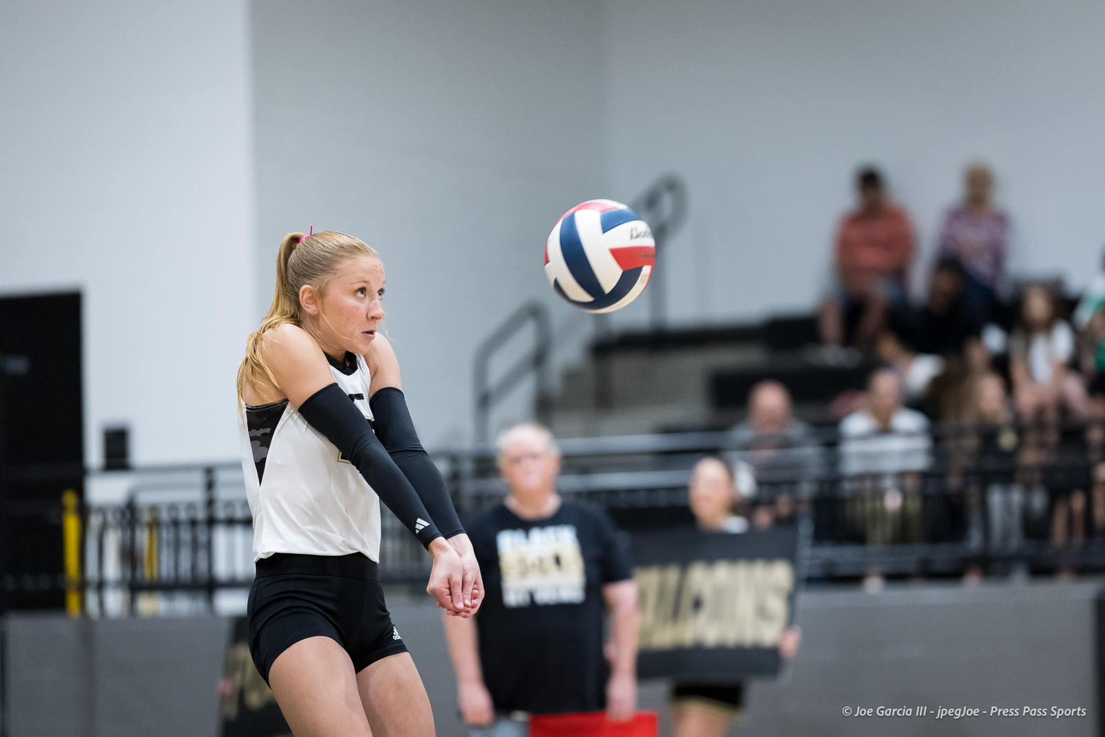 Female volleyball player in a white jersey and black arm sleeves prepares to contact the ball midair, with spectators in the background.