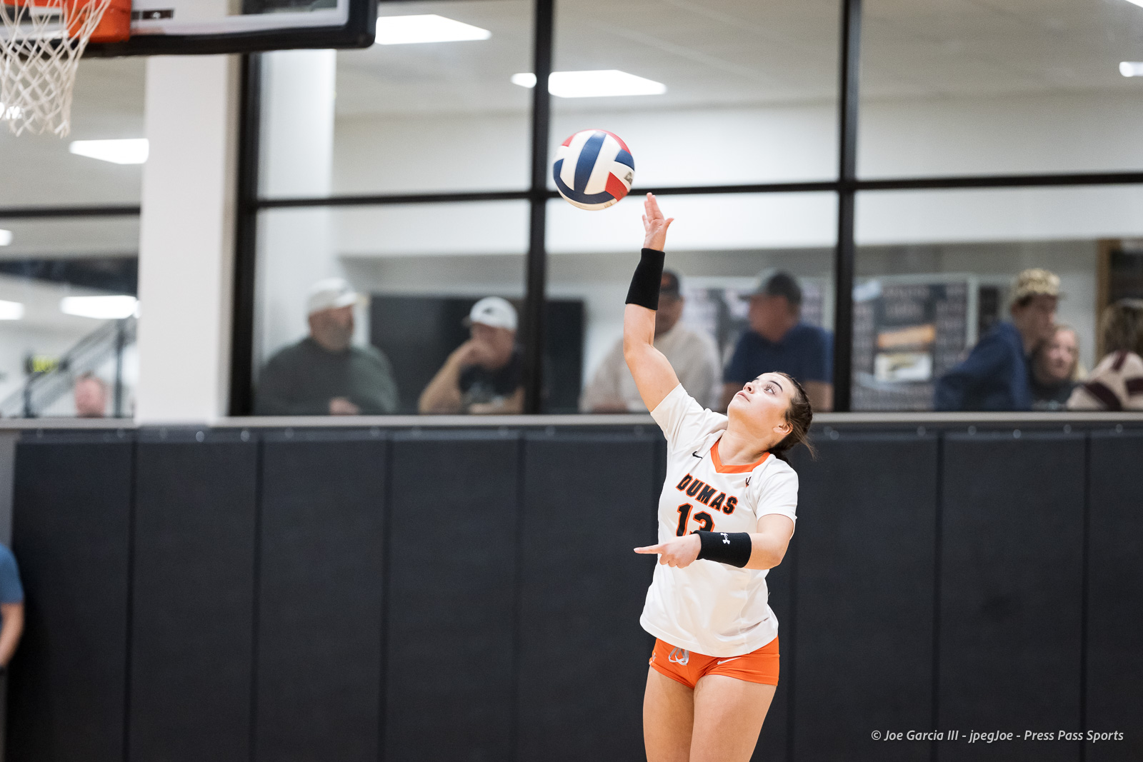 Female volleyball player in a white Dumas jersey (no. 13) raises her arm to hit the ball indoors, with spectators behind glass.